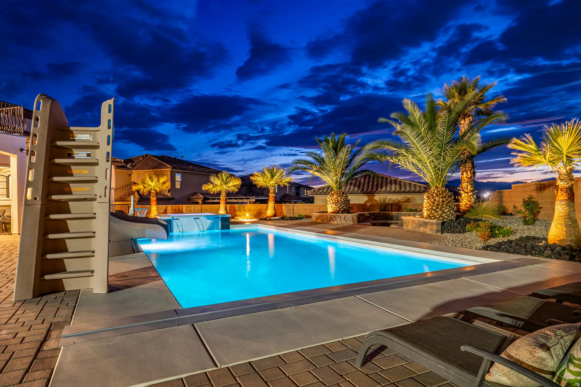 Illuminated swimming pool featuring a waterfall spa, integrated slide, paver patio, and mature palm trees