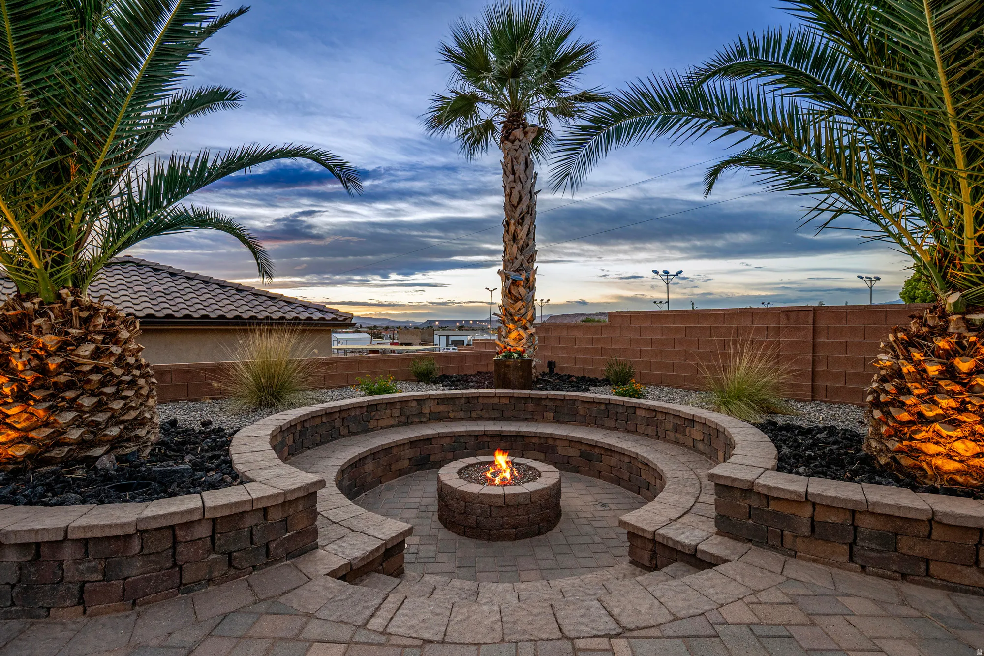 Paver patio featuring a circular stone fire pit with integrated stone seating, surrounded by mature palm trees