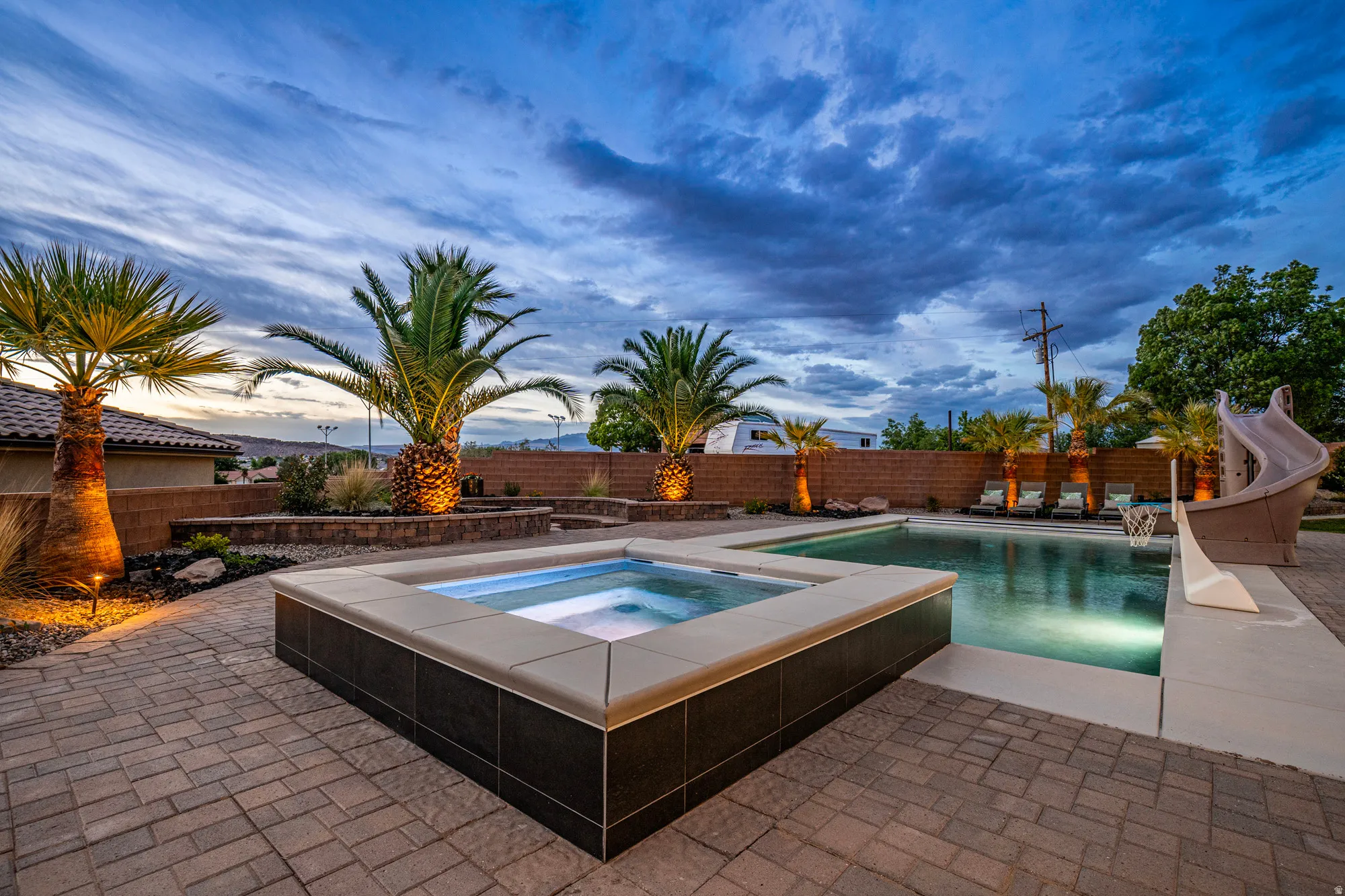 Outdoor oasis featuring a paver patio, integrated spa, swimming pool with a slide, and mature palm trees with accent lighting