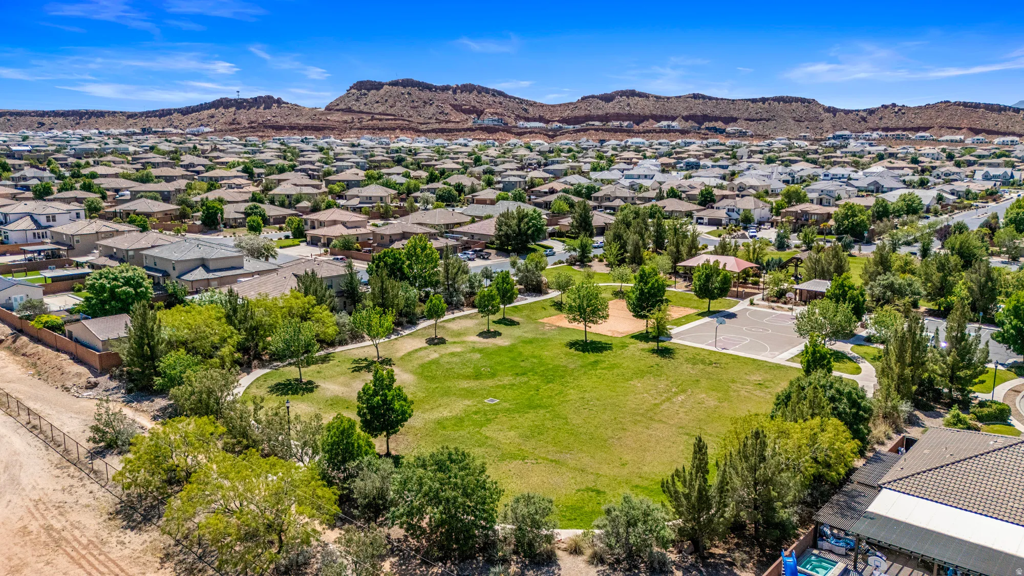 Expansive community park featuring a manicured green space, mature trees, paved walking paths, a basketball court, and shaded picnic areas