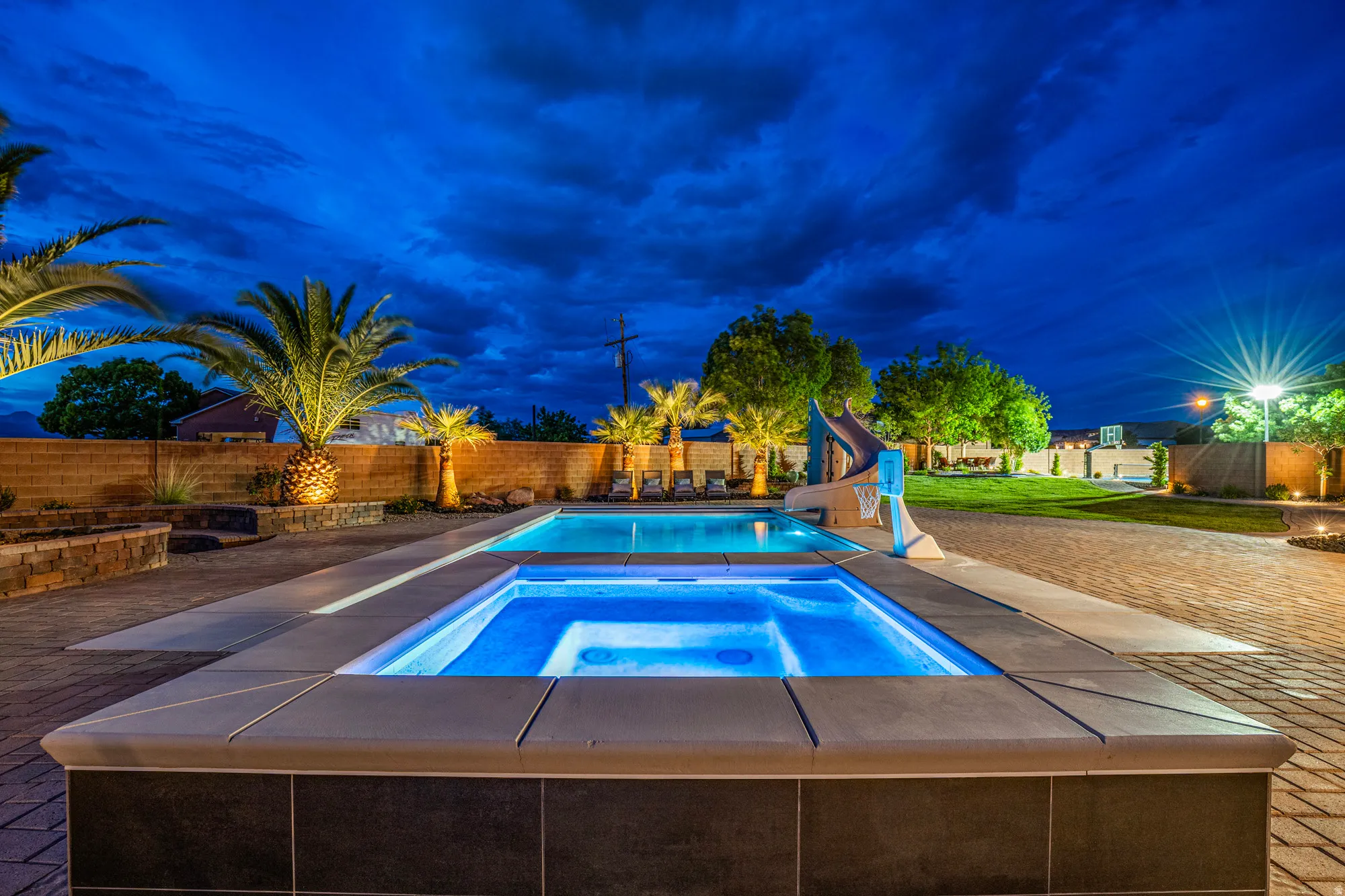Expansive outdoor living area featuring a blue-tiled spa, a rectangular swimming pool, and paver decking