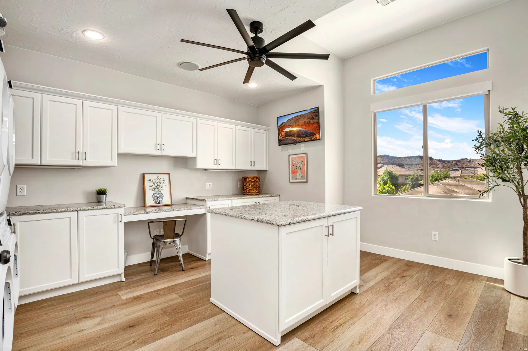 Bright room with wood-finish flooring featuring extensive white cabinetry, granite countertops, an integrated desk area, and a central island