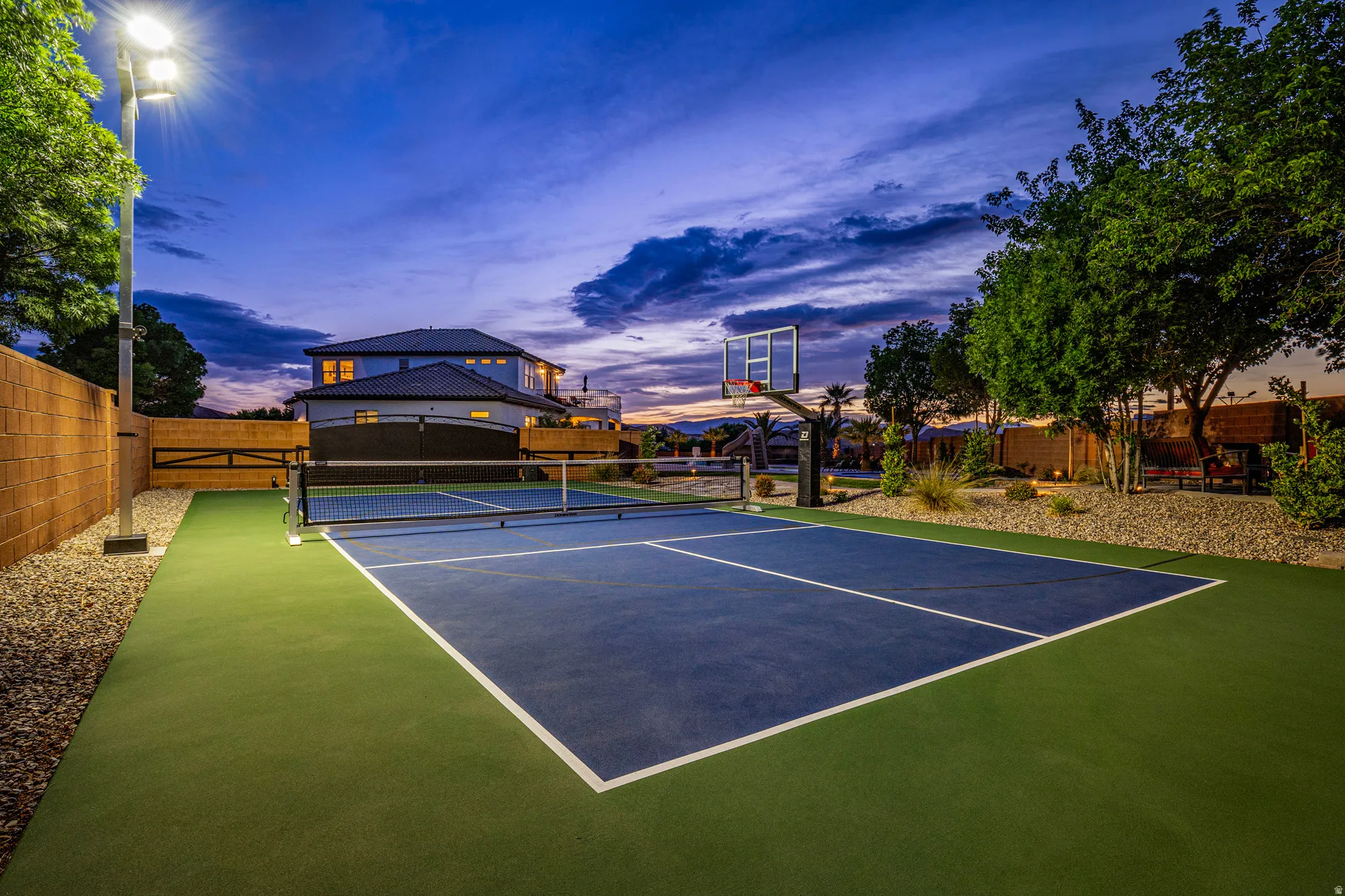 Outdoor multi-sport court featuring a blue playing surface with green perimeter, a basketball hoop, and a net