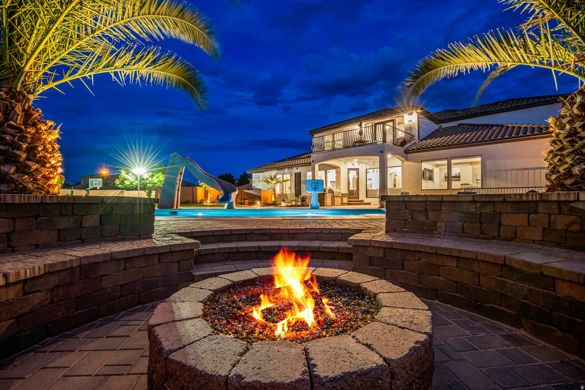 Expansive outdoor living area featuring a built-in fire pit with paver surround, integrated seating, and mature palm trees