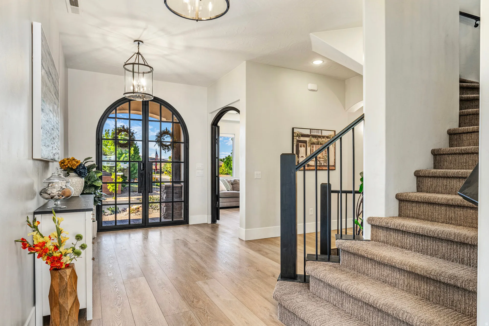 Arched double entry doors with grid pattern, light wood-finish flooring, staircase with carpeted treads and black metal railing, overhead chandelier lighting, and a white baseboard trim