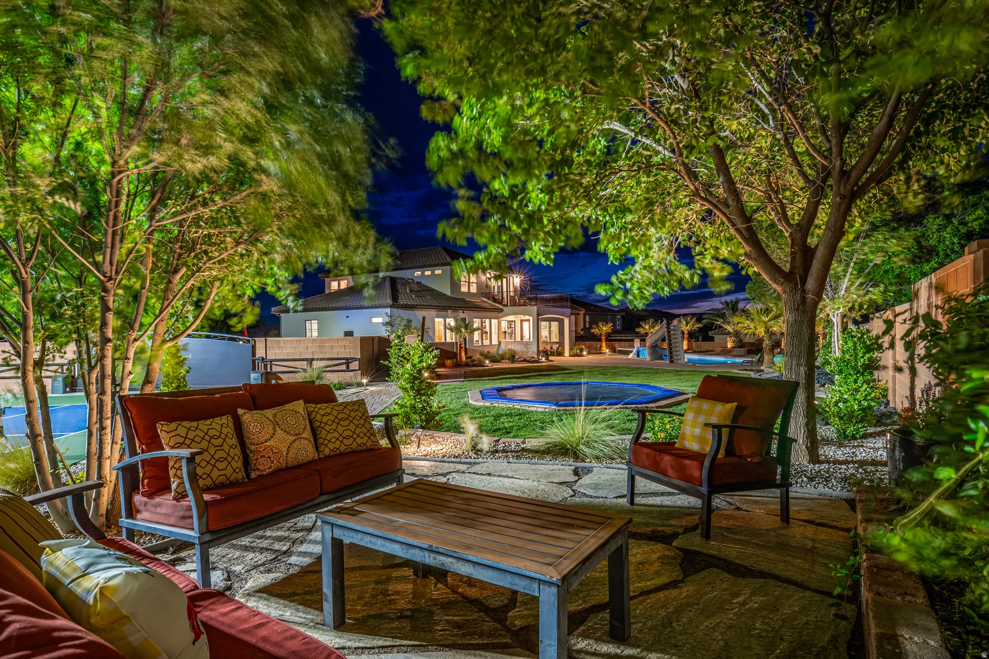 Expansive outdoor living area featuring a stone patio, mature trees, manicured lawn, and a covered swimming pool