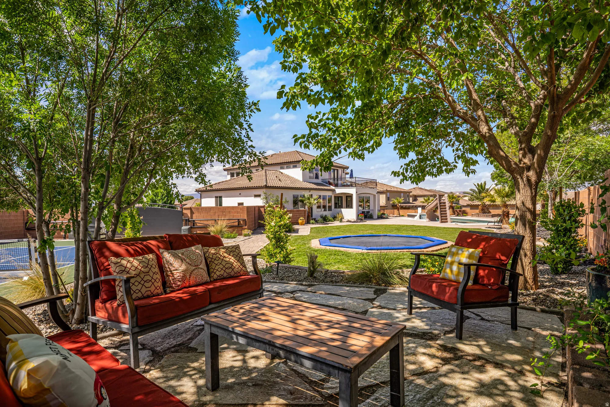 Expansive stone patio featuring mature shade trees, a manicured lawn, and a multi-level residence with a balcony