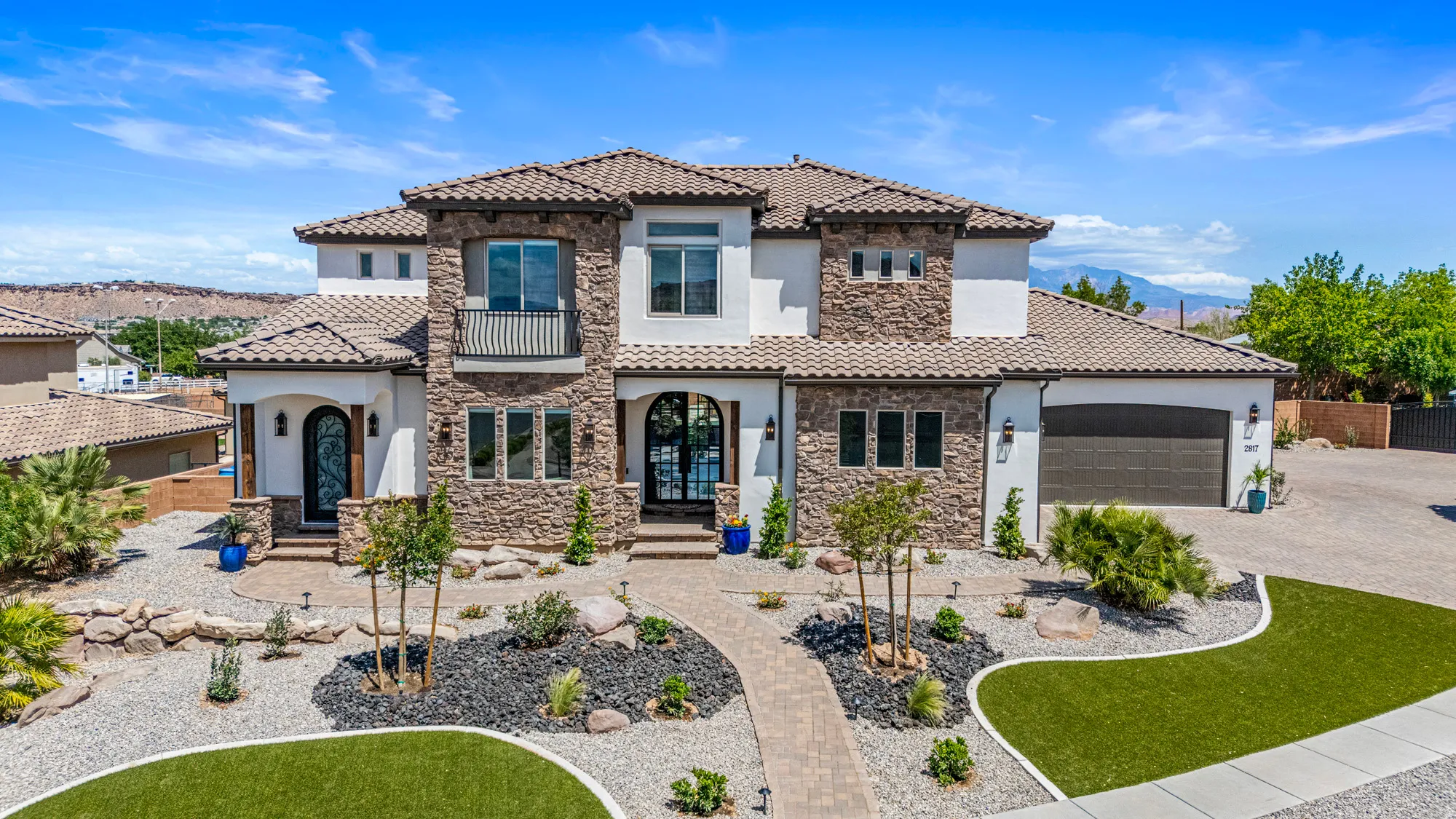 Stucco and stone exterior featuring a tile roof, arched entryways, a second-story balcony with iron railing, a two-car garage, and a paver pathway