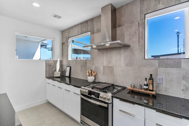 Modern kitchen featuring sleek white cabinetry, black stone countertops, a stainless steel range with a matching range hood, and a grey tile backsplash