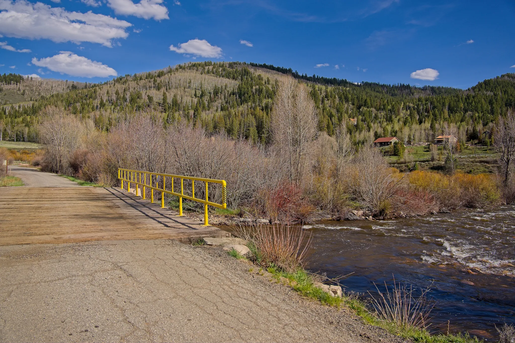 Rustic bridge with yellow railings crossing a flowing river, framed by natural foliage and a mountainous backdrop with distant structures