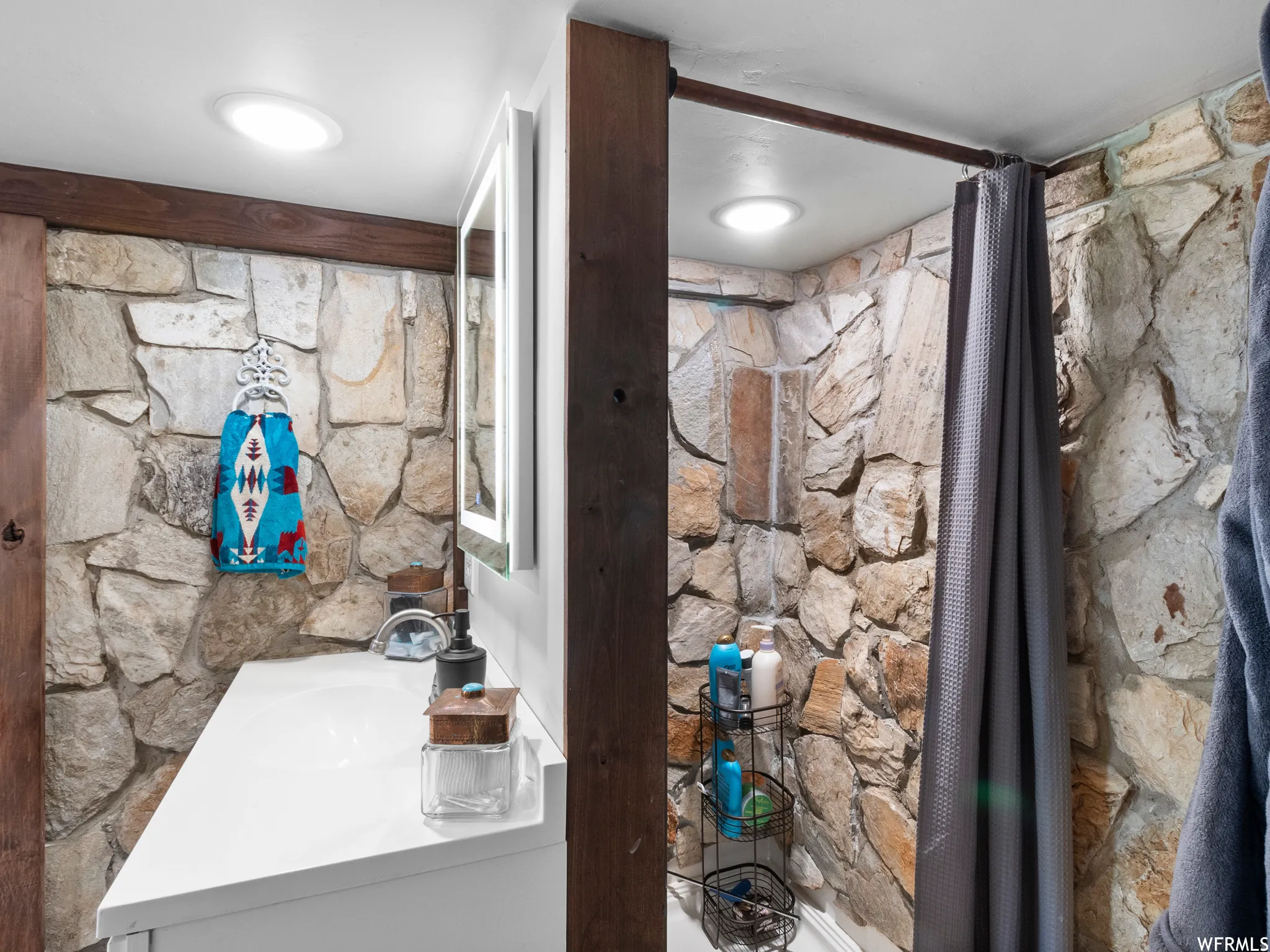 Bathroom featuring natural stone wall cladding, a white vanity with an integrated sink, an illuminated wall-mounted mirror, and recessed ceiling lighting