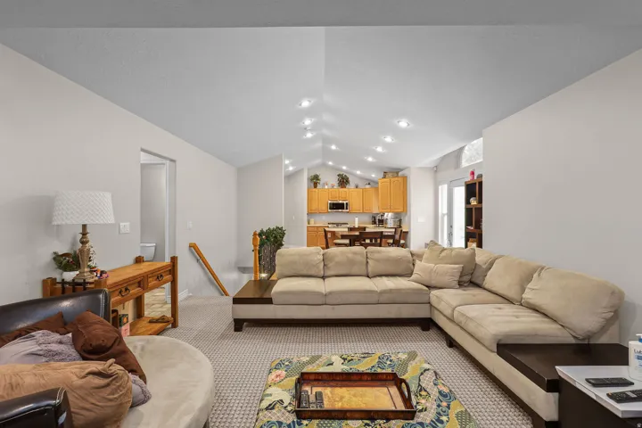 Carpeted living area featuring a vaulted ceiling with recessed lighting, a view into the kitchen with wood cabinetry, and a visible doorway to another area of the property