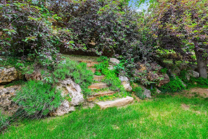 Landscaped yard featuring a stone staircase, natural rock formations, mature trees, and vibrant green foliage