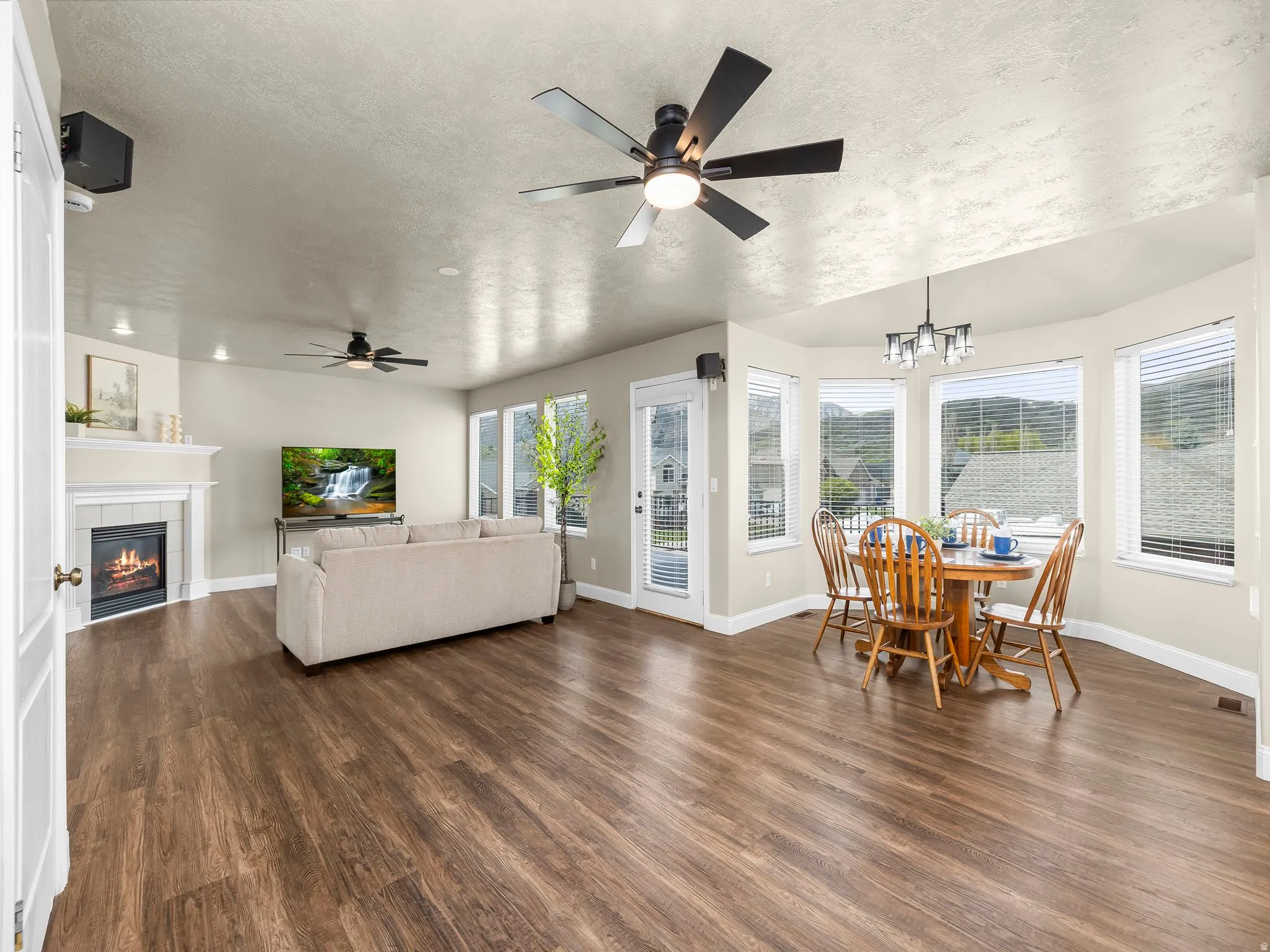 Spacious living area featuring wood-finish flooring, a fireplace with a white mantle, ceiling fans, and a dining nook with bay windows
