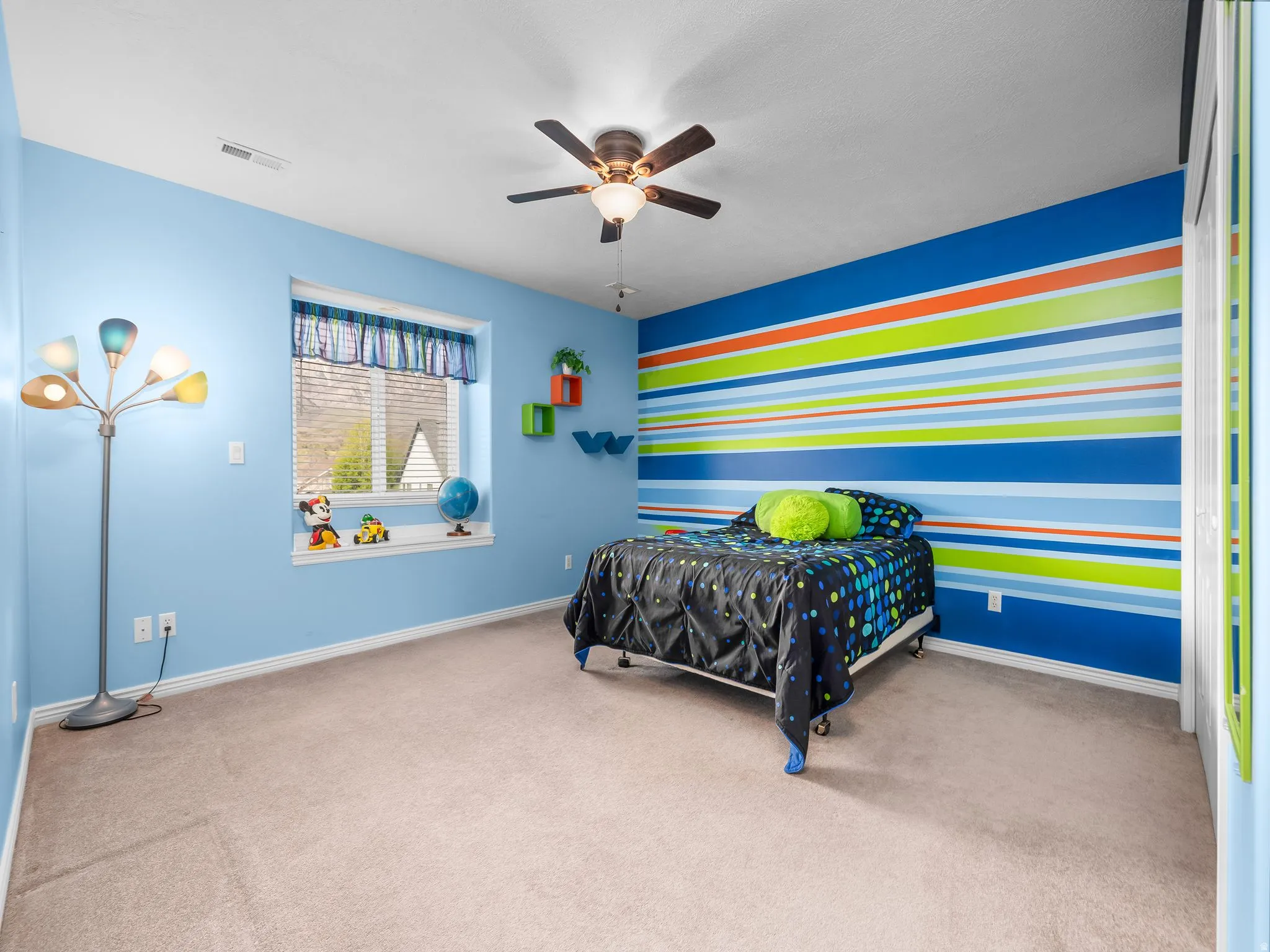 Carpeted room featuring a window with blinds, ceiling fan with integrated lighting, a colorful accent wall, and built-in shelving