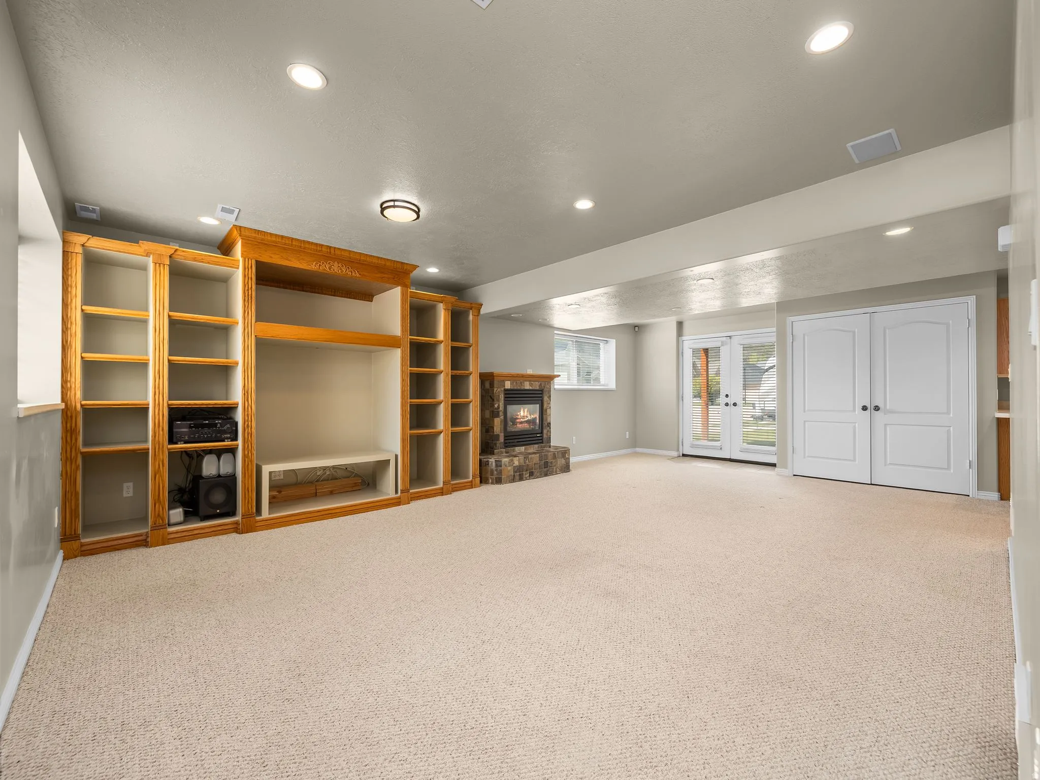 Expansive carpeted room featuring a stone-clad fireplace, built-in wood shelving, recessed lighting, and double glass-paneled doors with exterior access