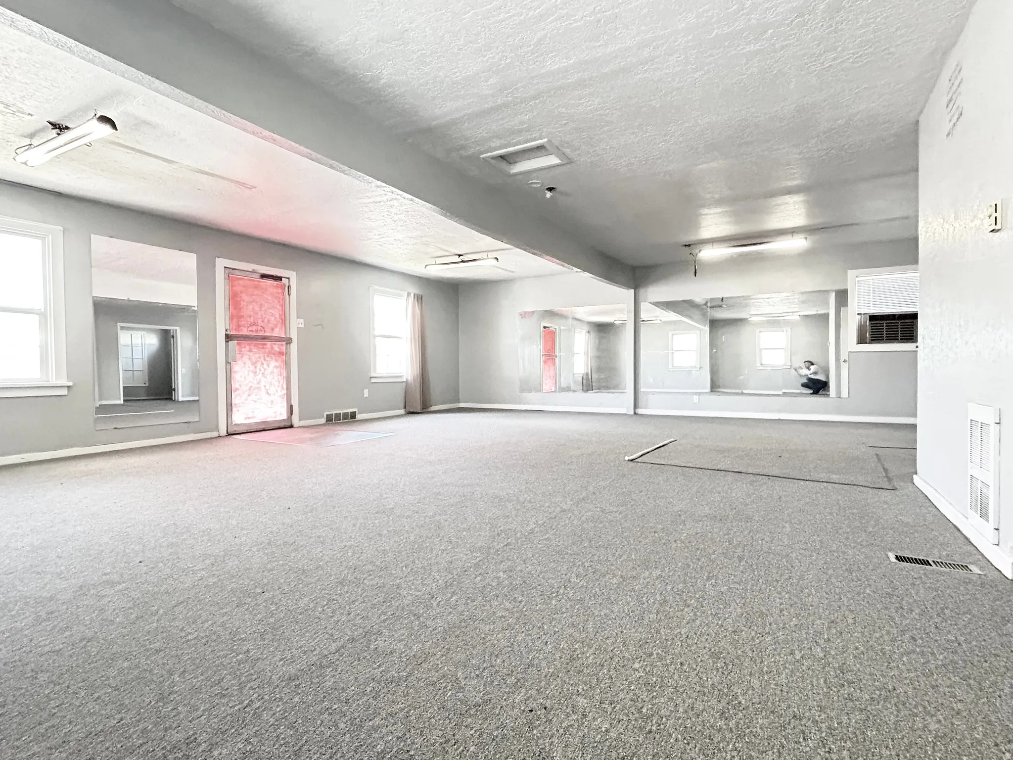 Spacious interior room featuring light gray carpeting, multiple windows, and extensive wall-mounted mirrors