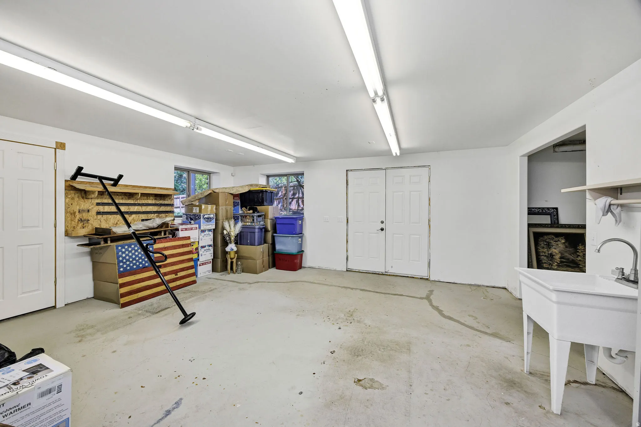 Spacious utility room featuring white walls, concrete flooring, multiple fluorescent lighting fixtures, and a utility sink with a chrome faucet