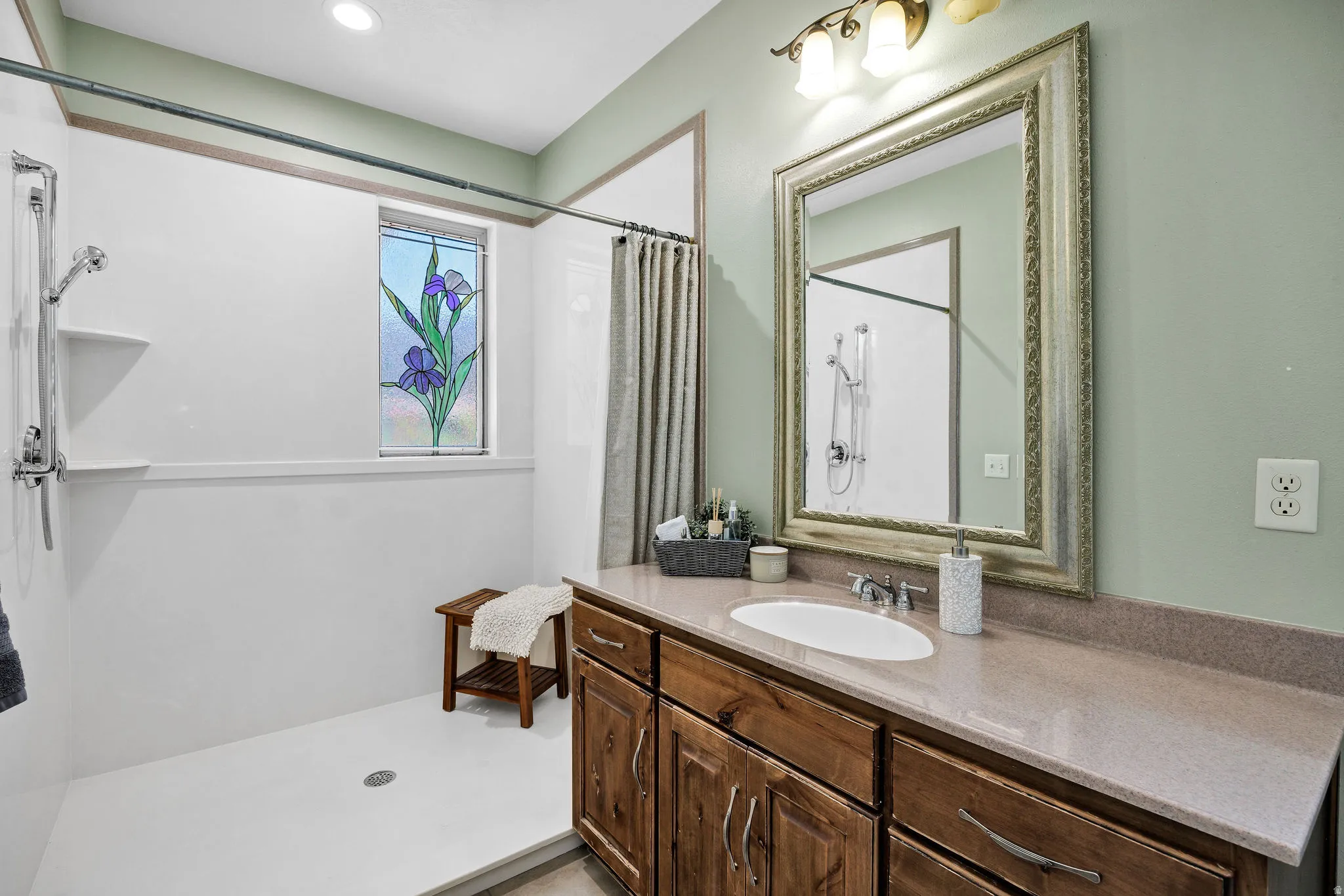 Bathroom featuring a single vanity with a solid surface countertop and undermount sink, framed mirror, and a walk-in shower with a decorative stained glass window
