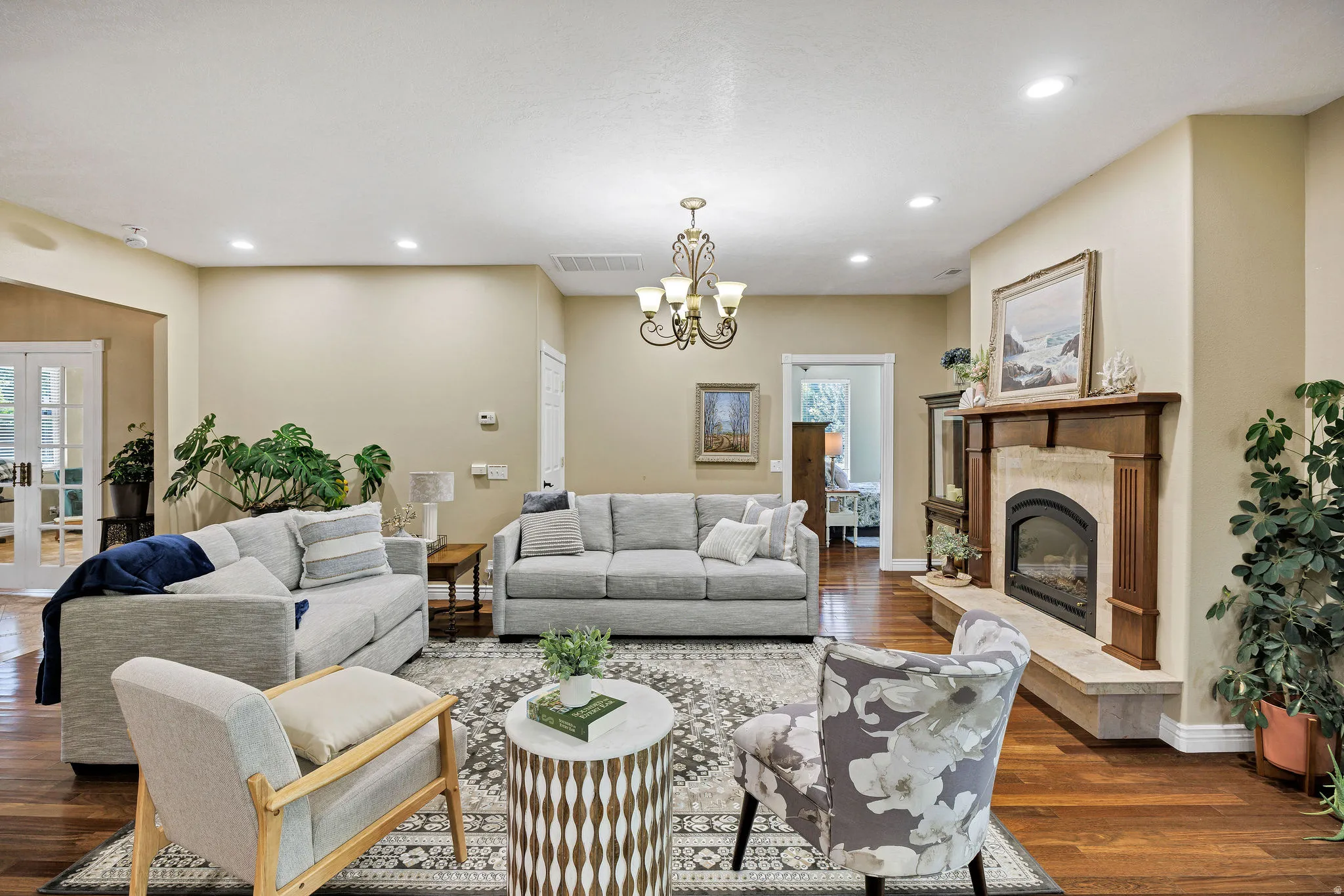 Spacious living area featuring rich wood-finish flooring, a decorative chandelier, recessed lighting, and a prominent fireplace with a wood mantle and stone surround