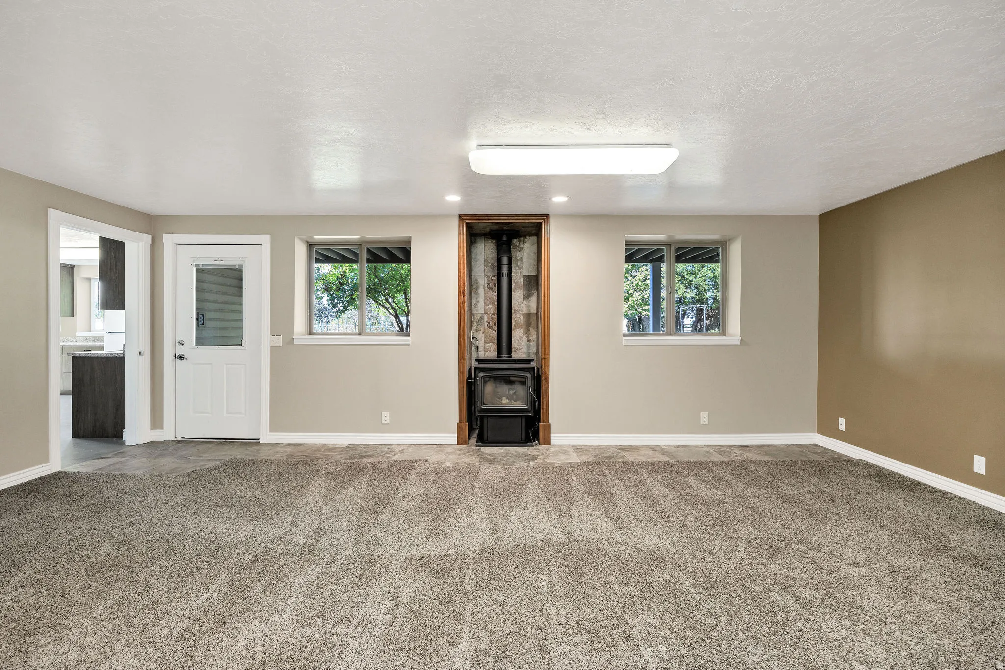 Spacious room featuring neutral paint, soft grey carpeting, a corner wood-burning stove, two windows, and a doorway leading to an additional space with wood-finish cabinetry