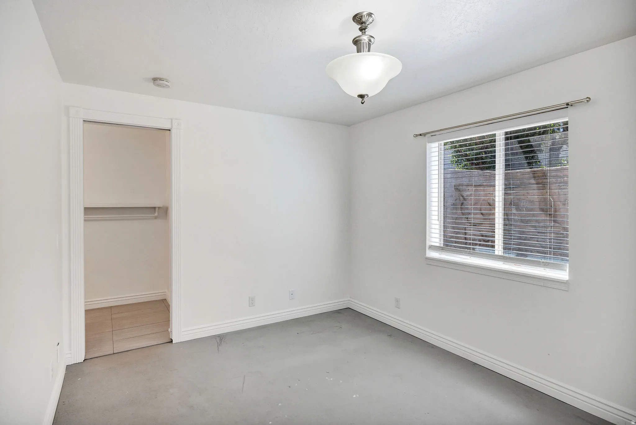 Room featuring a window with blinds, a ceiling-mounted light fixture, white walls, and a closet with wood-finish flooring