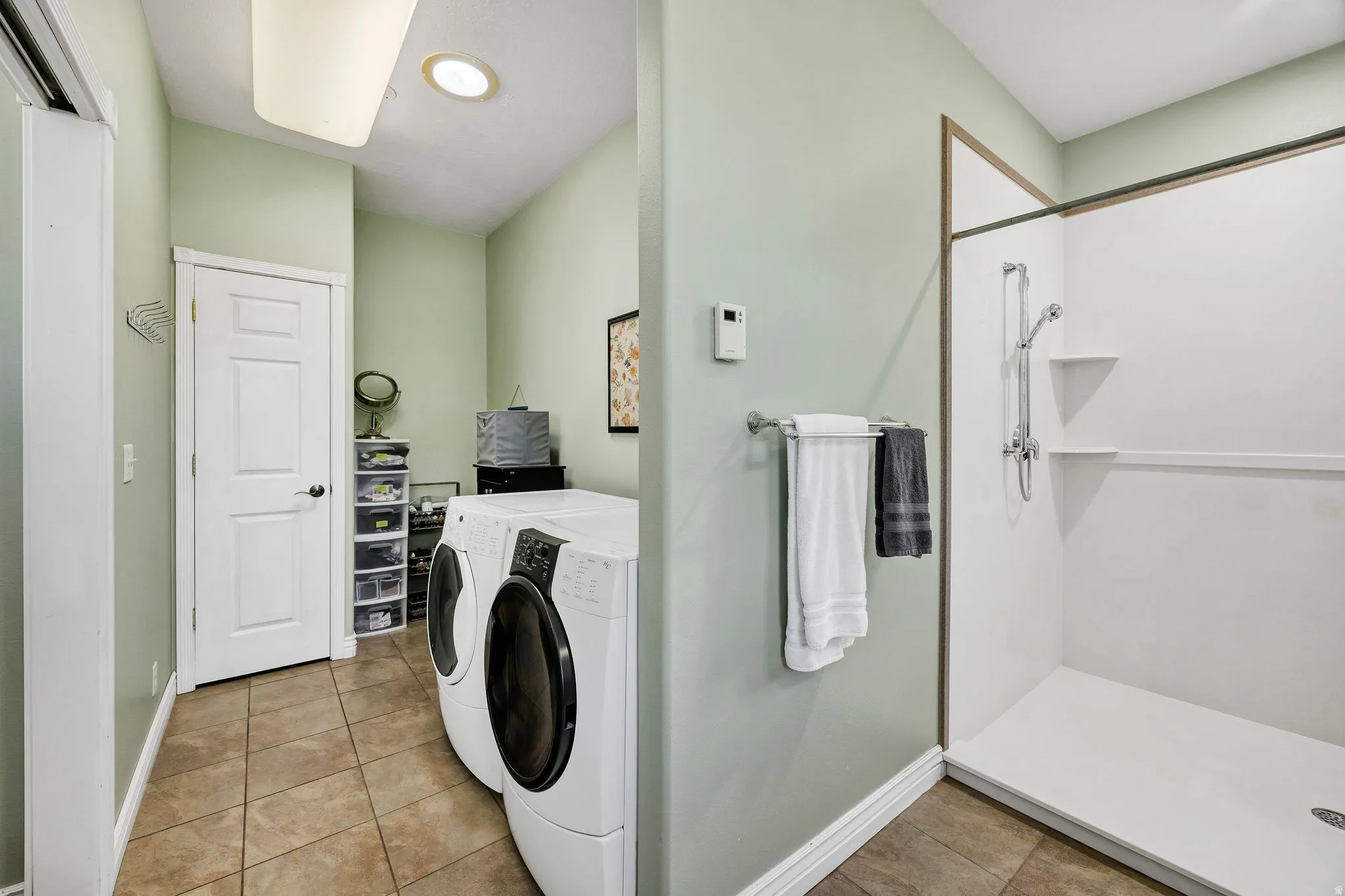 Combination laundry and shower room featuring tiled flooring, a white paneled door, a recessed ceiling light, and a white shower stall with built-in shelving