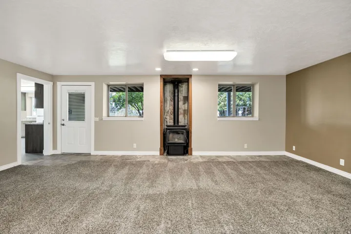 Spacious room featuring neutral paint, soft grey carpeting, a corner wood-burning stove, two windows, and a doorway leading to an additional space with wood-finish cabinetry