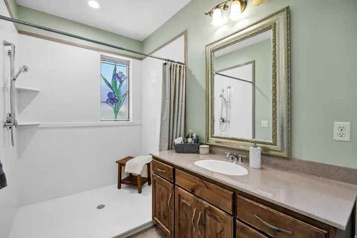Bathroom featuring a single vanity with a solid surface countertop and undermount sink, framed mirror, and a walk-in shower with a decorative stained glass window