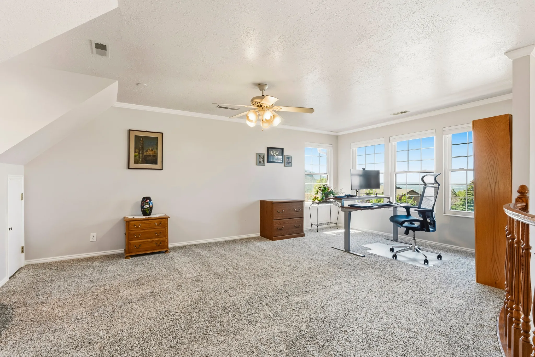 Spacious carpeted room featuring multiple windows, a ceiling fan, crown molding, and a partial view of a wood-finish staircase railing