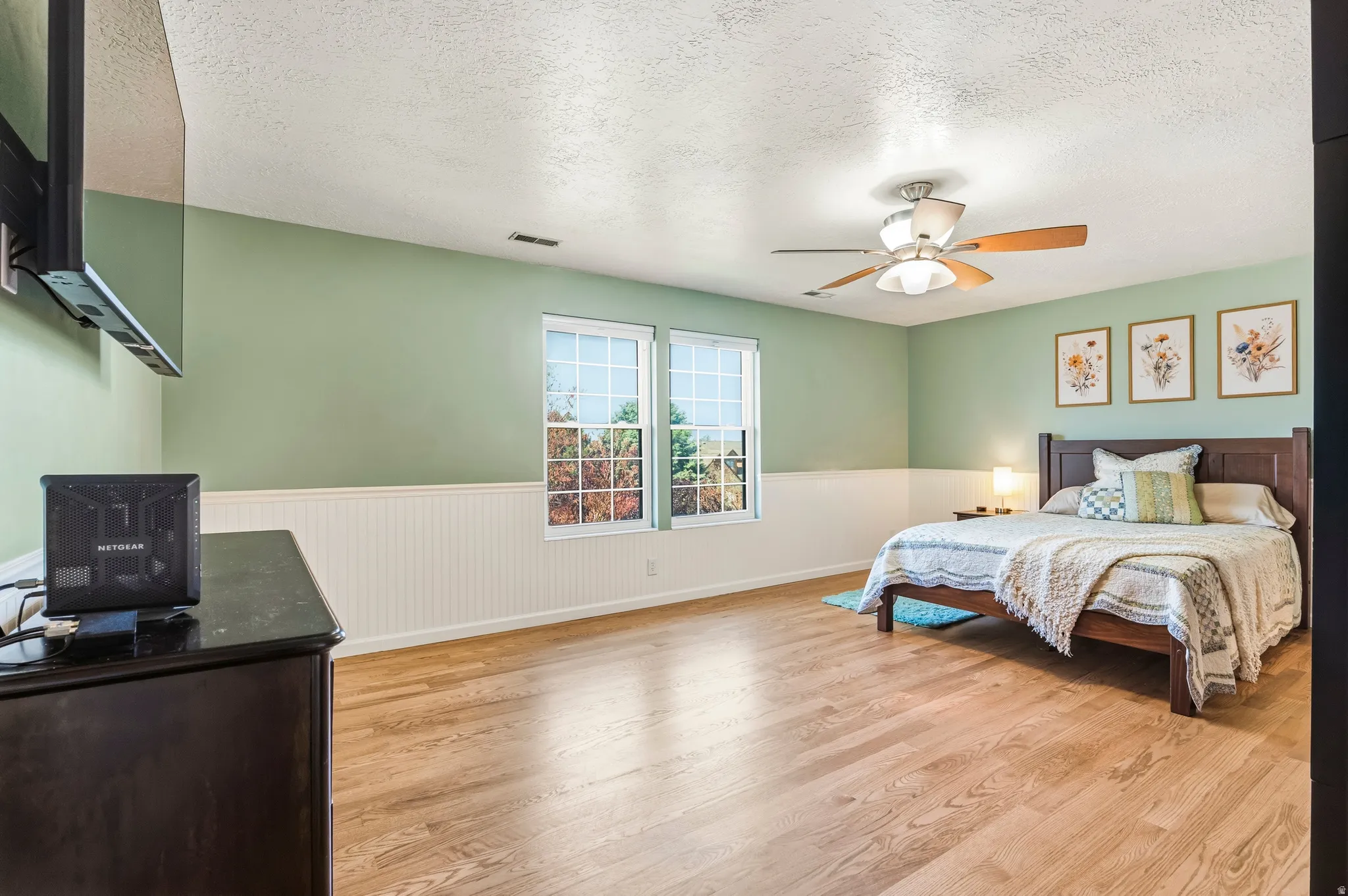 Spacious room featuring wood-finish flooring, two large windows, white wainscoting, a ceiling fan, and ample wall space