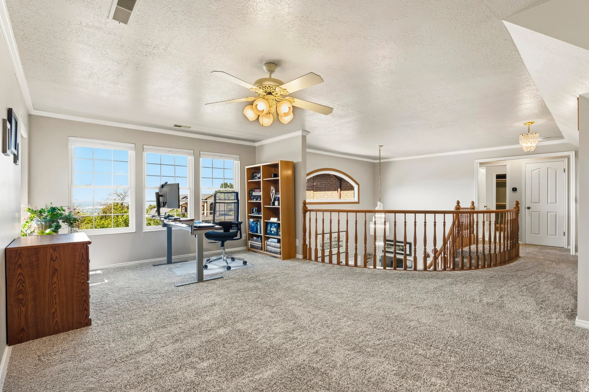 Carpeted loft area featuring a curved wood banister, three large windows, a built-in wood bookshelf, and a ceiling fan light fixture