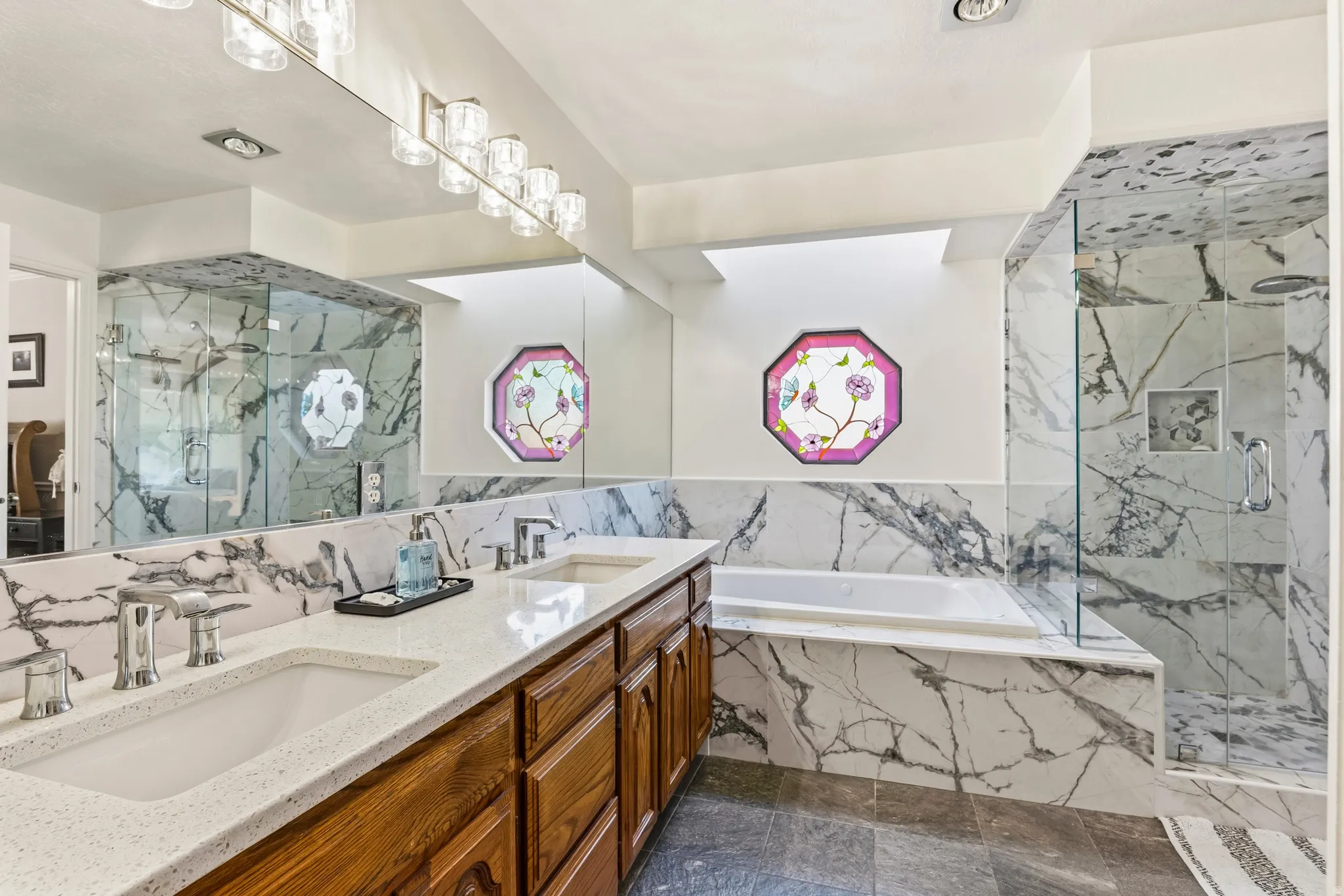 Spacious bathroom featuring a dual vanity with white countertops, a built-in soaking tub, a glass-enclosed shower, marbled wall tiling, and a decorative octagonal window