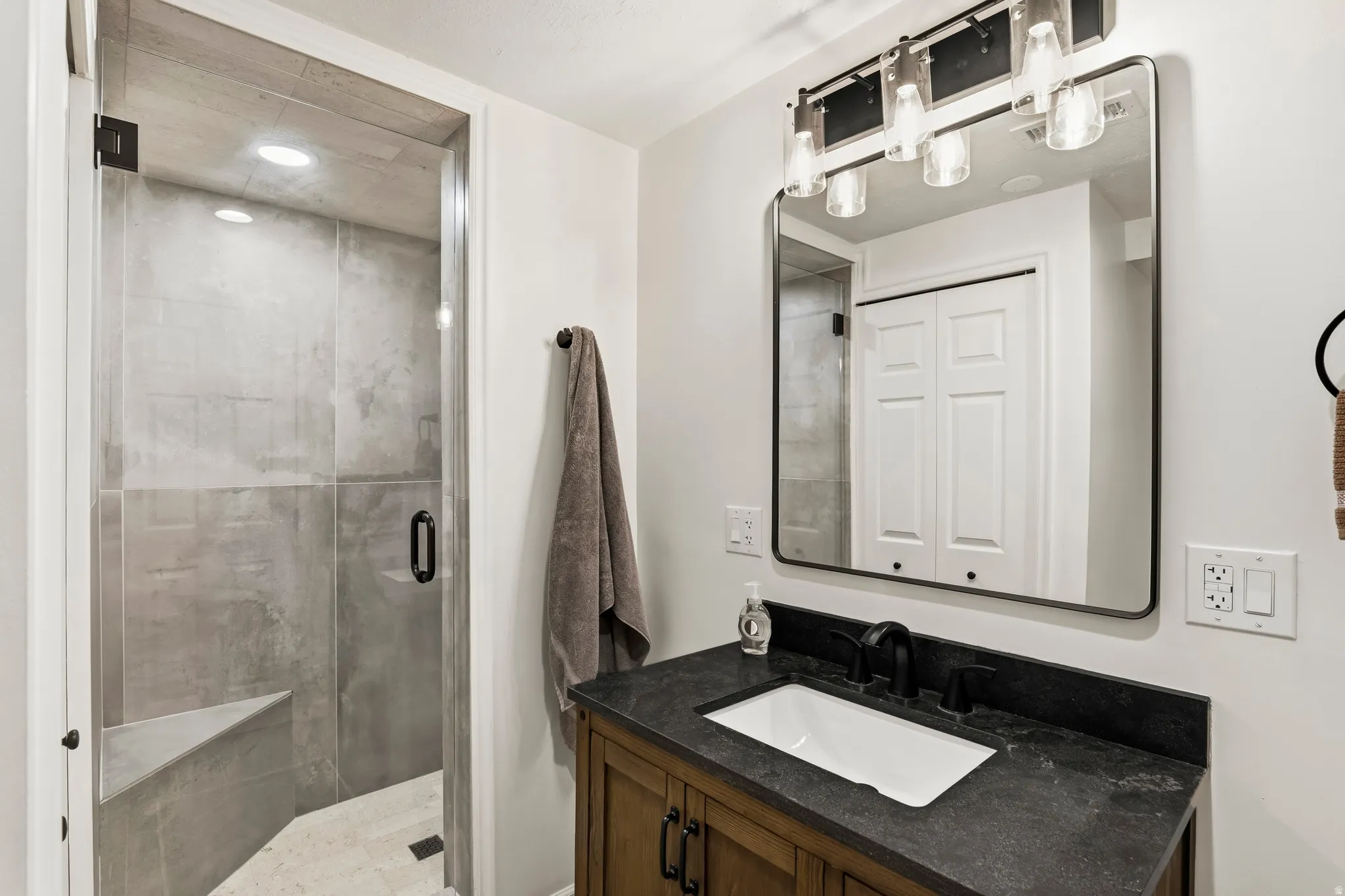 Bathroom featuring a glass-enclosed shower with a built-in bench, wood-finish vanity with an undermount sink, dark stone countertop, and matte black fixtures