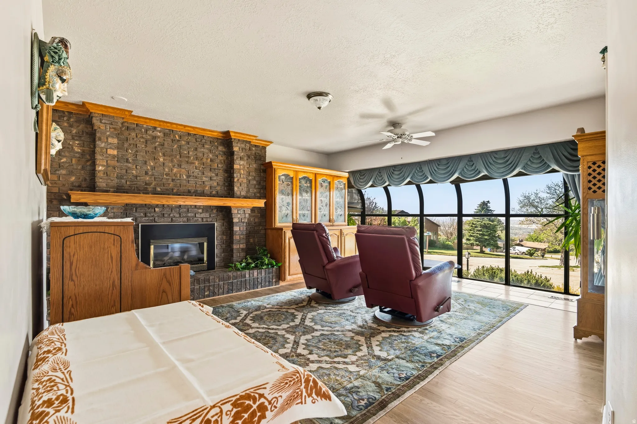 Living area featuring a floor-to-ceiling curved window wall, a brick-surround fireplace with a wood mantle, wood-finish flooring, a ceiling fan, and built-in wood cabinetry