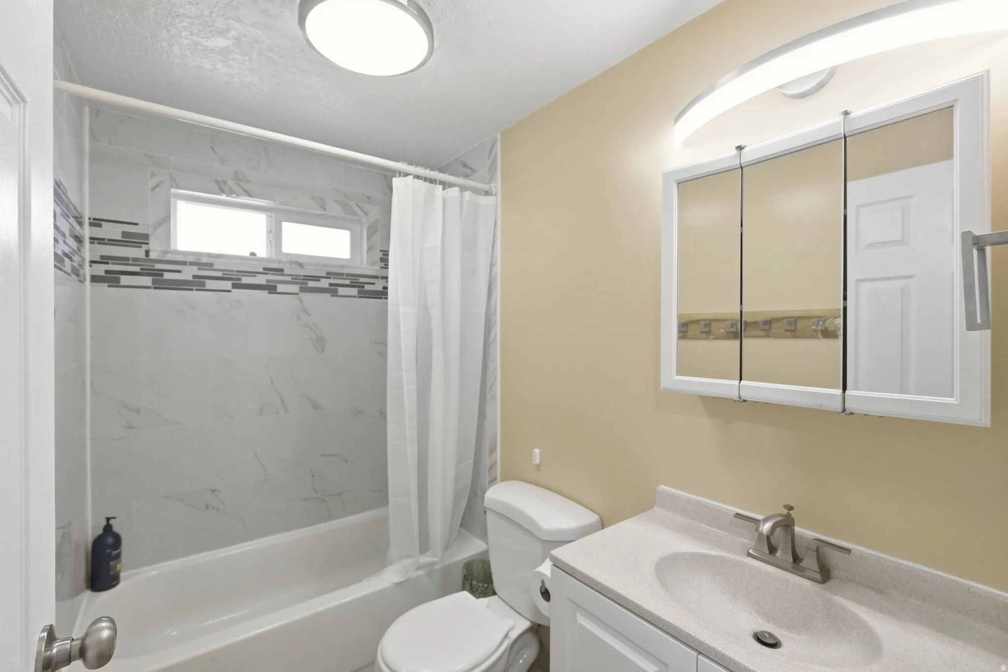Bathroom featuring a bathtub with subway tile surround and decorative accent band, a dual-pane horizontal window, a white vanity with integrated sink, a mirrored medicine cabinet, and a wall-mounted light fixture