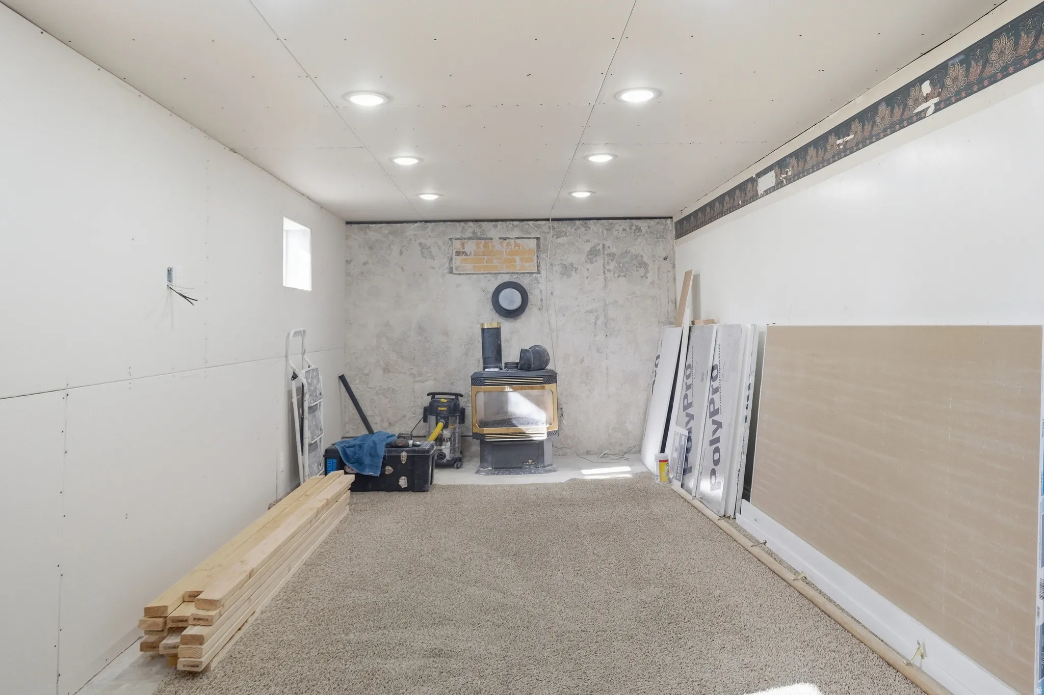 Recessed ceiling lighting illuminating a carpeted room featuring a window, partial drywall installation, and a wood-burning stove