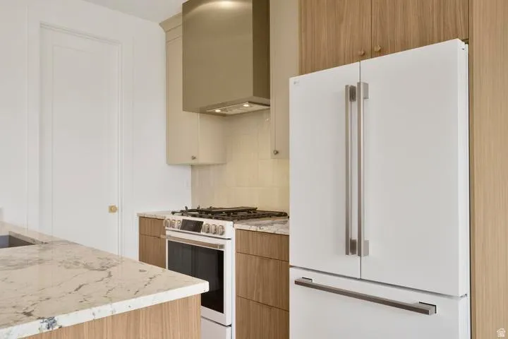 Kitchen featuring a white French-door refrigerator, stainless steel range with matching hood, light-toned wood-finish cabinetry, and veined stone countertops