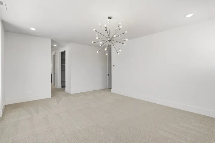 Spacious room featuring a contemporary sputnik chandelier, recessed lighting, neutral carpet flooring, crisp white walls, and a visible doorway to a hallway with built-in shelving