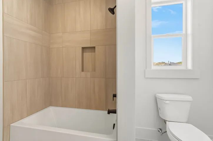 Full-size bathtub with surrounding light wood-finish wall tiles, featuring a built-in shower niche and matte black fixtures