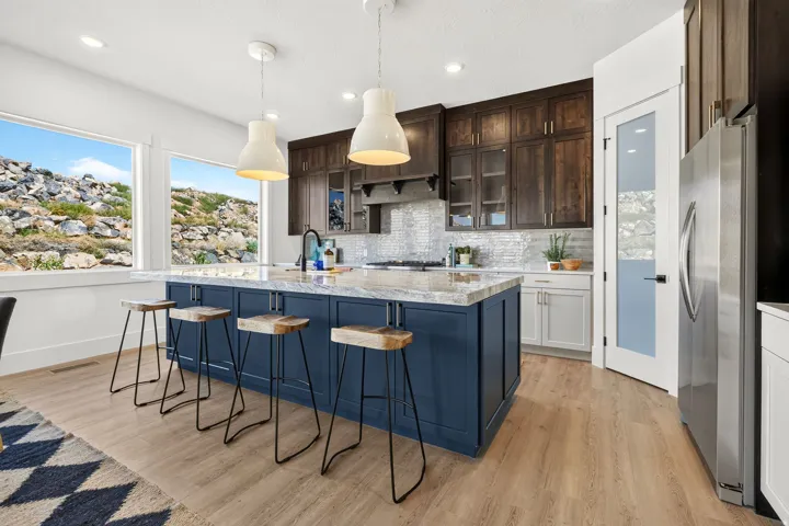 Spacious kitchen featuring a dark blue island with a light-colored countertop, light wood-finish flooring, two large pendant lights, dark wood cabinetry, and a stainless steel refrigerator