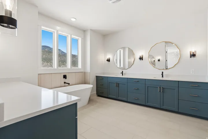 Contemporary bathroom featuring a freestanding soaking tub, triple windows with a mountain backdrop, a dual vanity with teal cabinetry and white countertops, and decorative round mirrors with sconce lighting