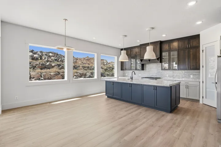 Open-concept kitchen featuring a spacious island with sink, light wood-finish flooring, dark wood cabinetry with glass accents, and a light-toned tile backsplash