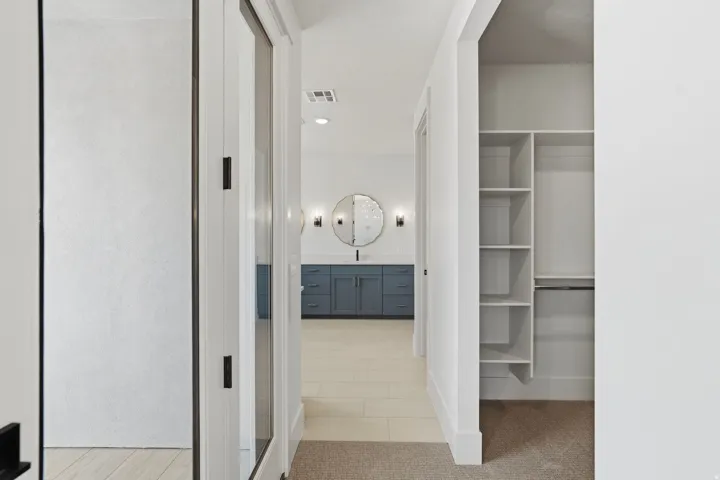 Hallway featuring a built-in closet with shelving and hanging rods, leading to a bathroom with a dual vanity, round mirror, and wall sconces