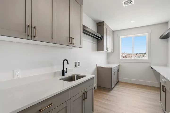 Dedicated laundry room featuring light gray shaker cabinetry, expansive white countertops, a built-in stainless steel sink with a matte black gooseneck faucet, and light wood-finish flooring