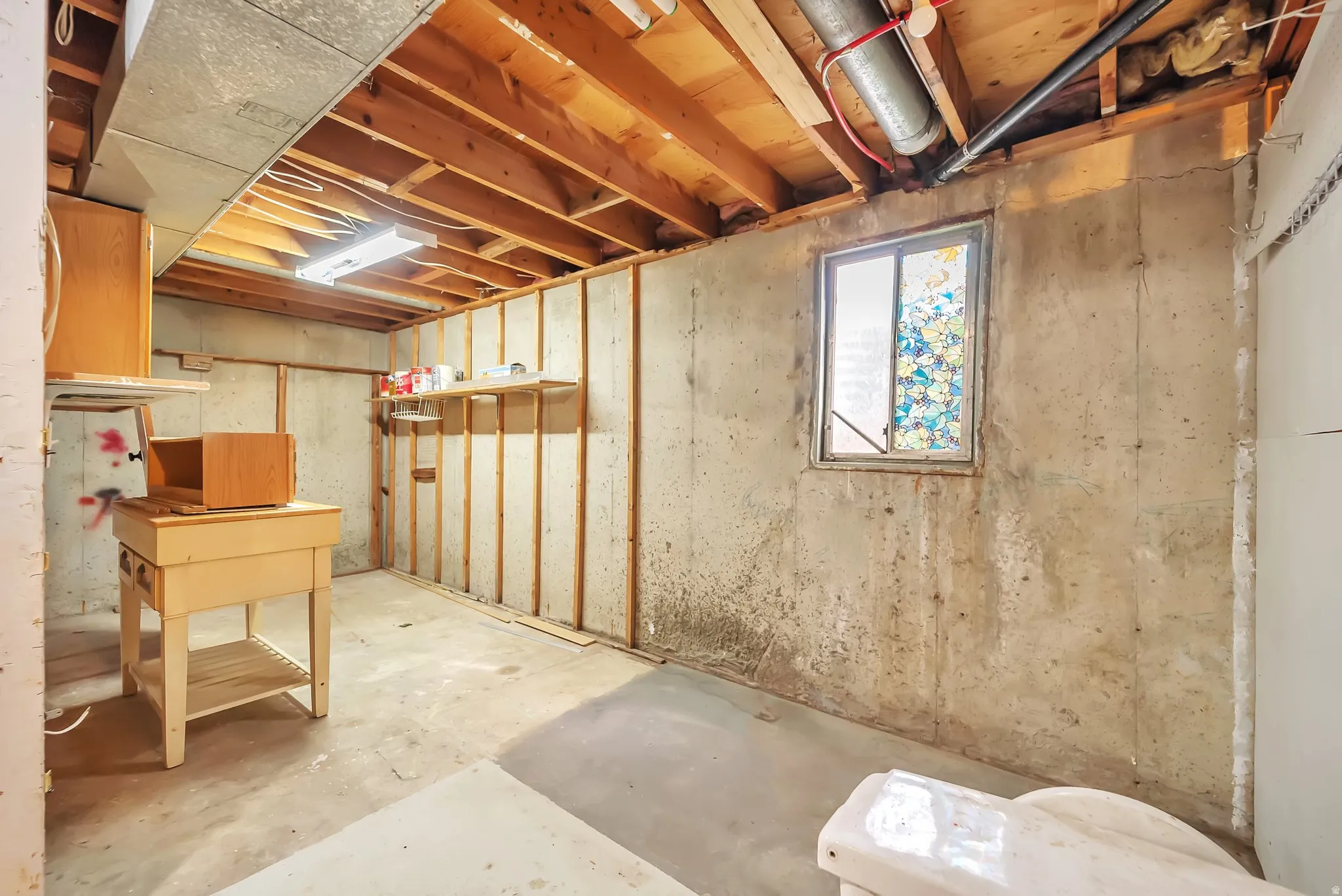 Unfinished basement space featuring exposed wooden ceiling joists, concrete flooring, a single window, and visible plumbing and ductwork