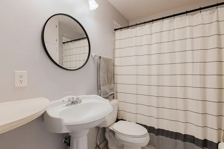 Pedestal sink with a dual-handle chrome faucet, round black-framed mirror, and wall-mounted light fixture