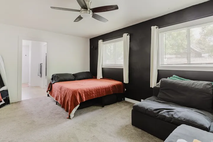 Carpeted room featuring a ceiling fan, two windows with blinds, a dark accent wall, and a doorway opening to an additional area