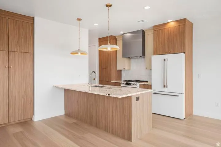 Kitchen featuring an island with a light-toned countertop, wood-finish cabinetry, stainless steel appliances, pendant lighting, and light wood-finish flooring