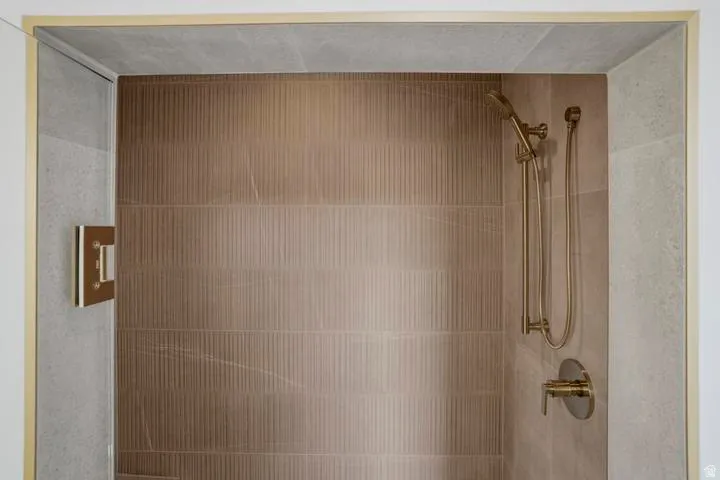 Shower enclosure featuring light grey stone-look tile, fluted brown-toned wall tile, a brass-finish shower system, and brass-finish hardware